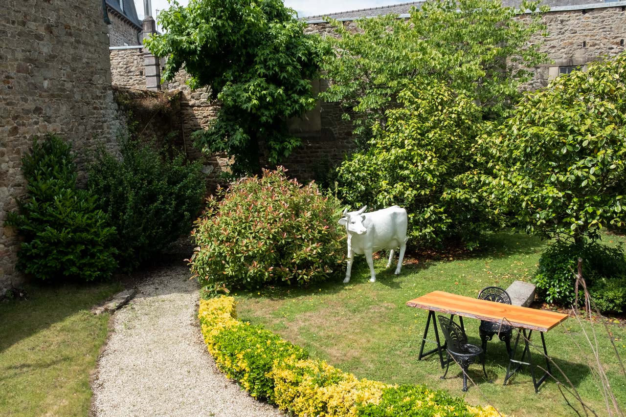 Vu du jardin de la demeure. Chemin en graviers, table avec deux chaises et sculpture d'une vache.